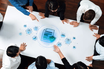 A diverse business team collaborates around a large table, analyzing documents and digital data with icons illustrating technology and security, highlighting teamwork in a professional setting. Trope