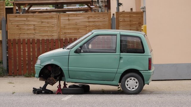 Green car with missing front wheel on an urban roadside, supported by a jack. The scene includes overcast lighting, wood fencing, and a pavement setting.