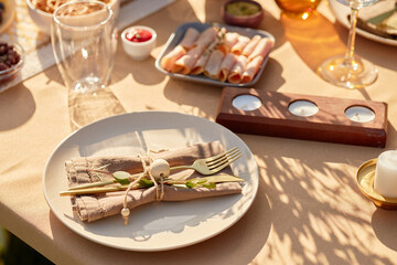 Table setting featuring empty plate with neatly arranged fork and knife on linen napkin, surrounded by appetizers and candles, sunlight casting shadows across surface