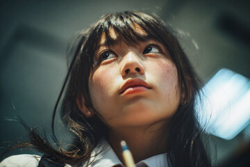 Close-up portrait of a pensive schoolgirl holding a pencil and looking upward under soft classroom lighting
