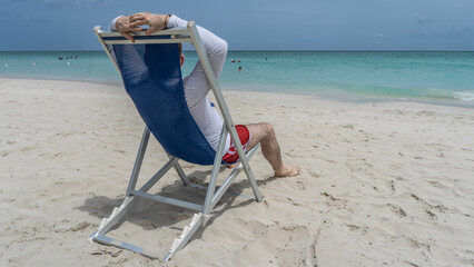 A man is sitting on a chaise longue on a sandy beach, looking at the ocean. Hands behind your head. Silhouettes of people in the turquoise sea. The blue sky.  Cuba. Varadero. Resort. Hotel.  