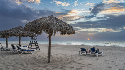 Sunset by the ocean. Two people are lying on deck chairs, a glass with a drink in the hand. Straw...