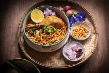 Beautiful still life of traditional Northern Thai Khao Soi chicken curry noodles served in an enamel bowl on a wooden table, with crispy noodles, shallots, lime, chili oil.