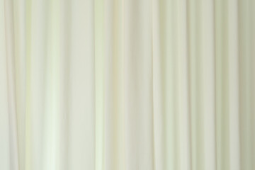 Fototapeta premium Background of a creamy white or light beige fabric curtain. The curtain hangs in gentle, vertical folds, creating a smooth, flowing texture. The lighting is diffused, giving the fabric a subtle
