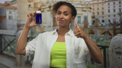 Woman holding blue bottle with raised hand and thumbs up gesture in front of ancient building and stone ruins while smiling; health confidence endorsement.