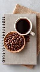 Overhead View Of A Flat Lay Coffee Scene Featuring Roasted Coffee Beans In A Small Bowl Next To A White Mug Filled With Dark Coffee Placed On A Textured Notebook And Burlap Fabric