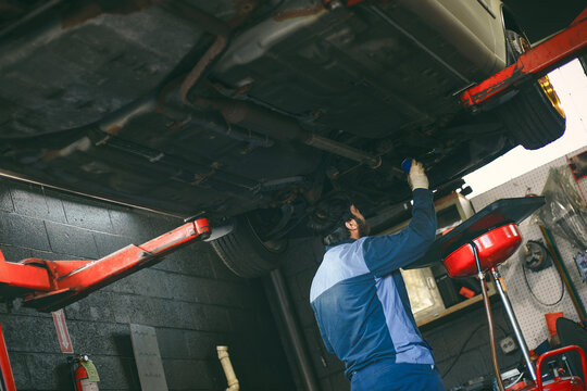 A man in overalls stands in a car repair shop against the backdrop of a car being repaired.