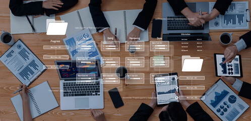 A high-angle view of a business meeting showcasing professionals engaged in discussion with laptops and tablets. Charts and documents indicate strategic planning and collaboration efforts. Trope