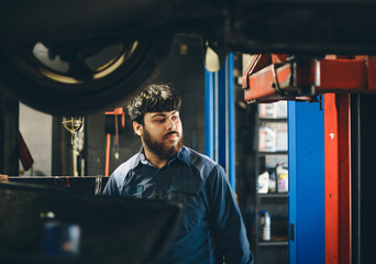 A man in overalls stands in a car repair shop against the backdrop of a car being repaired.