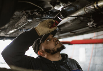 A mechanic holds a flashlight while inspecting the underside of a car in a garage. The environment suggests a typical automotive repair setting.