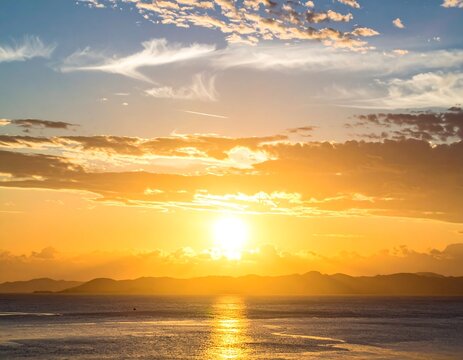 Golden hour paints the sky above the ocean, reflecting light on the water, with mountains in the distant horizon. The clouds add texture