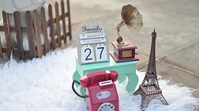 An adorable baby crawling on a soft white rug in an outdoor photoshoot setup, surrounded by decorative props such as a vintage telephone, wooden fence, stuffed toy, Eiffel Tower model, and date calend