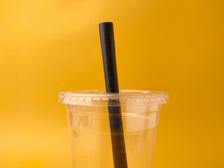 The image shows a clear disposable plastic cup with a lid and a black straw on a yellow background