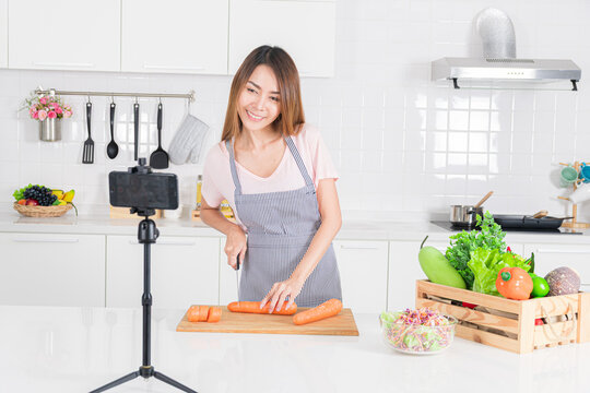 Young asian woman chopping carrot while filming a cooking demonstration in her bright white kitchen emphasizing healthy eating.