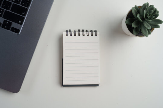 A small spiral notebook placed beside a laptop and a green plant on a white table, creating a calm and minimal workspace mood.