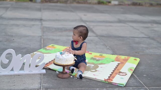 A cute baby celebrating a first birthday with a cake smash during an outdoor photoshoot. The child is sitting on a colorful play mat next to a decorated birthday cake and a &ldquo;One&rdquo; sign, wearing a cute 