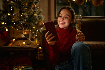 Happy woman using smart phone while drinking champagne on Christmas at home.