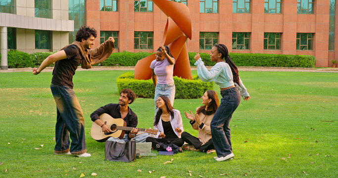 Indian young adult gen z male man hold guitar play new music lady woman sing song have fun joy. Happy pupil girl boy guy group spend day break time do funny dance enjoy relax mood at garden park place