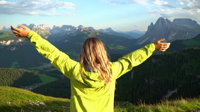 Young caucasian woman with long blonde hair in yellow windbreaker raises arms on Dolomites viewpoint at golden hour, slow crane up & dolly out. Concept of freedom & serenity