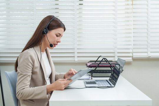 Asian businesswoman adult female operator using tablet during online meeting, headset on, multitasking while consulting customer and managing communication support process in modern corporate office