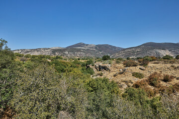 Naklejka premium Mountainous countryside filled with lush green and dry brush, showing mixed vegetation, boulders, and layered hills in the distance