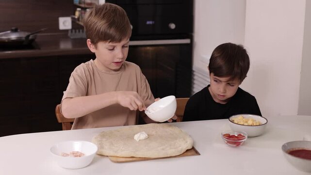 Children preparing pizza at home and spreading white sauce on dough, concept of family cooking, learning and creativity.