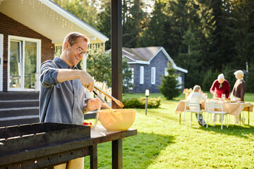 Caucasian middle aged man grilling food outdoors using barbecue grill while smiling, senior...