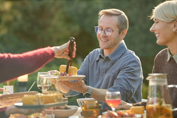 Caucasian middle aged man smiling while receiving grilled corn from another during outdoor meal with friends, sitting at table with food and drinks, enjoying social gathering