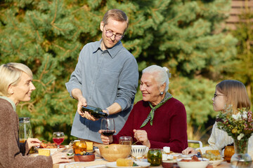 Caucasian middle aged man pouring wine for senior Caucasian woman while sitting at outdoor table, with woman and child enjoying meal together