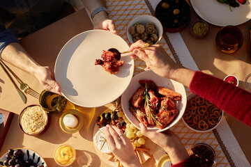 Senior woman serving chicken wings from bowl onto plate man holding, surrounded by assorted snacks...