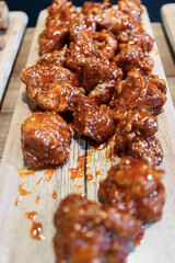 Vertical close-up of Crispy Korean Fried Chicken (Dakgangjeong) pieces coated in a glossy, sweet, and sticky red sauce. Served on a light wooden board. Popular K-Food street snack and comfort food.