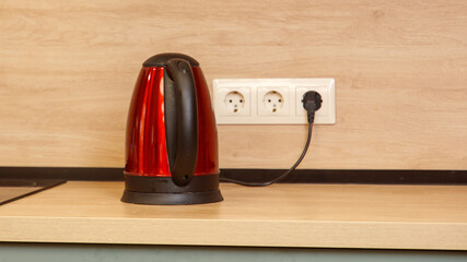 A red electric kettle sits on a wooden counter next to a power outlet