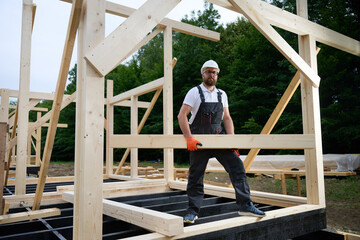 Construction worker installing wooden beam at house frame construction site. Carpenter in safety gear building timber structure for new home outdoors.