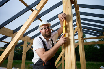 Construction worker using spirit level to check vertical alignment of wooden beam at frame house site. Builder wearing hard hat and safety glasses working on wooden structure outdoors.