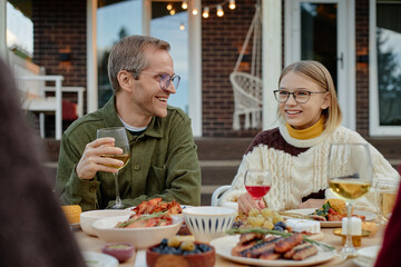 Caucasian middle aged man and his daughter sitting outdoors at table sharing meal and drinks with family, smiling and talking, surrounded by plates of food and beverages