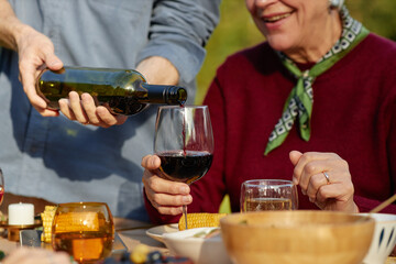 Senior Caucasian woman smiling while holding wine glass, young adult Caucasian man pouring red wine outdoors, enjoying meal together at table with food and drinks