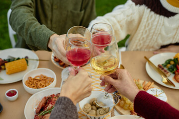 Four people clinking glasses during outdoor meal, hands visible above table with assorted food,...