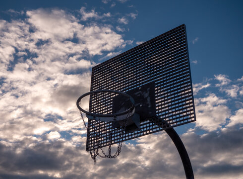 Outdoor basketball hoop with metal chain net against a dramatic cloudy sky. Low-angle view of street basketball basket in open air.