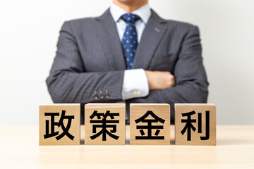 Obraz premium 「政策金利」と書かれたブロックと腕組みをするビジネスマン