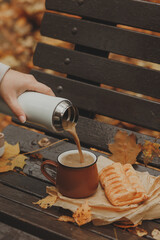 On the bench, there is a brown mug filled with hot coffee from a white thermos. A fresh bun is placed nearby, creating a cozy and homey atmosphere.