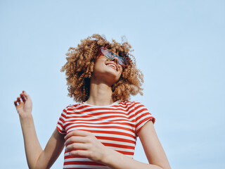 Obraz na płótnie Canvas Young person with curly hair smiles under a clear blue sky, wearing a red and white striped shirt and sunglasses, radiating carefree summer energy and playful confidence. Obraz na płótnie Canvas Young person with curly hair smiles under a clear blue sky, wearing a red and white striped shirt and sunglasses, radiating carefree summer energy and playful confidence.