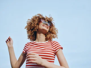 Fototapete Rund Musik Young person with curly hair smiles under a clear blue sky, wearing a red and white striped shirt and sunglasses, radiating carefree summer energy and playful confidence.  © SHOTPRIME STUDIO