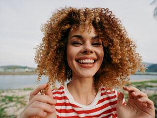 Joyful portrait of a young woman with curly hair smiling broadly outdoors by a lake, wearing a red...