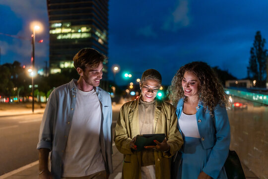 Diverse friends walking city street sharing tablet at night