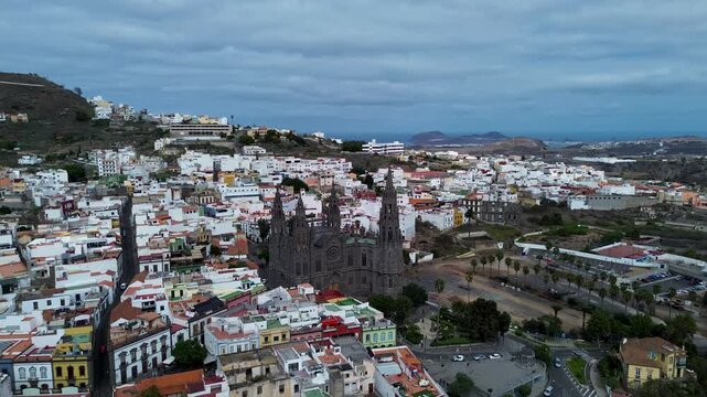 aerial view of the town of Arucas, Gran Canaria, featuring the prominent, dark Gothic Revival Church of San Juan Bautista rising above the dense cluster of white and colored traditional buildings.