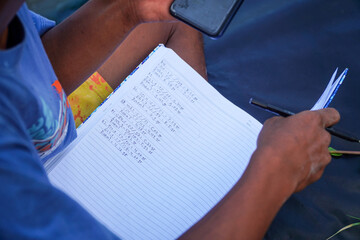 Overhead capture presents a person taking notes on a notebook while referring to a mobile phone, engaged in calculations and data recording for documentation and analysis purposes.