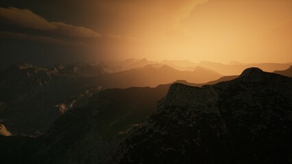 Golden hues blanket the landscape as the sun sets behind towering mountains, creating a stunning contrast of light and shadow. A serene atmosphere envelops the majestic scene.