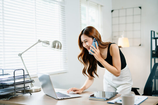 Smiling businesswoman talking on phone while working on laptop in bright modern office, symbolizing success, communication
