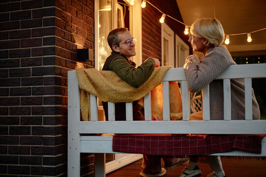 Middle aged Caucasian man and middle aged Caucasian woman sitting on porch bench talking and smiling at each other during evening, string lights hanging above, cozy outdoor setting - Powered by Adobe