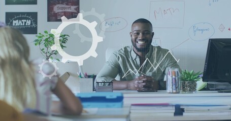 Smiling male teacher explaining math formulas at desk in classroom, with whiteboard and notebooks
